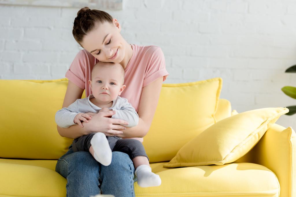 νεαρή μητέρα που αγκαλιάζει τον μικρό γιο ενώ κάθεται στον καναπέ - Φωτογραφία, εικόνα