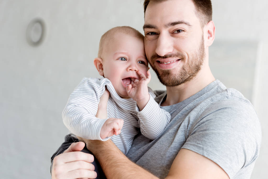 młody ojciec uśmiechający się trzyma małego chłopca w domu   - Zdjęcie, obraz