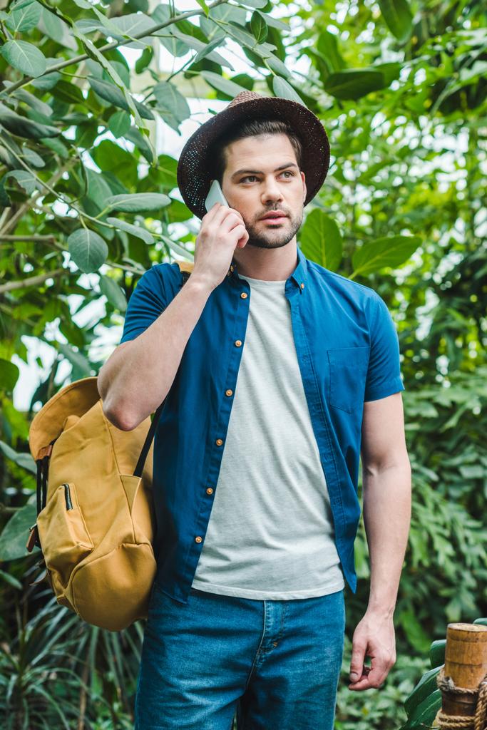 pohledný mladý muž ve stylovém oblečení mluví po telefonu v lese - Fotografie, Obrázek