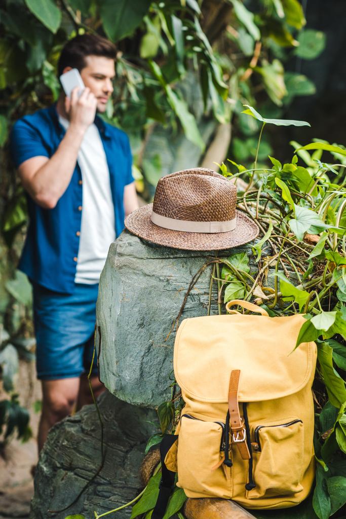 красивий молодий чоловік розмовляє по телефону в тропічних лісах з рюкзаком і солом'яним капелюхом на передньому плані
 - Фото, зображення