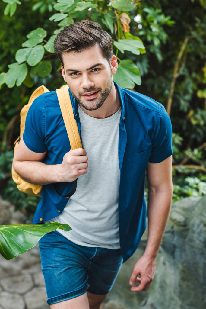 smiling young man in stylish clothing with backpack walking in park - Photo, Image