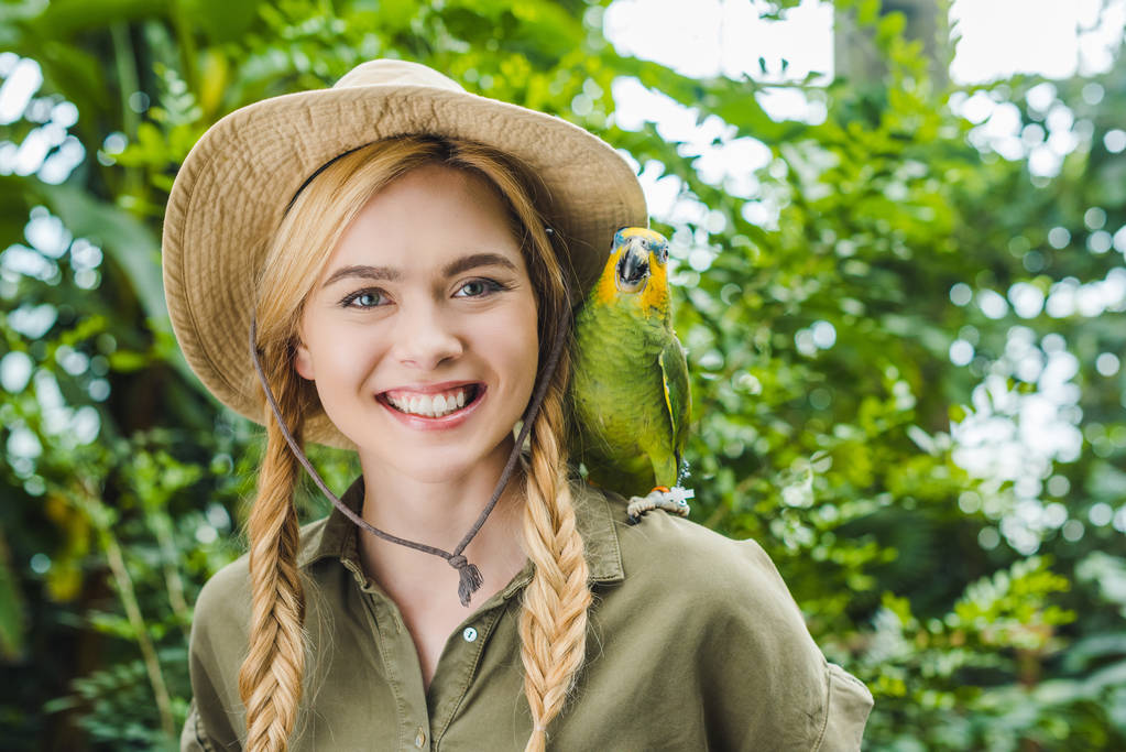 glückliche junge Frau im Safarianzug mit Papagei auf der Schulter im Dschungel - Foto, Bild