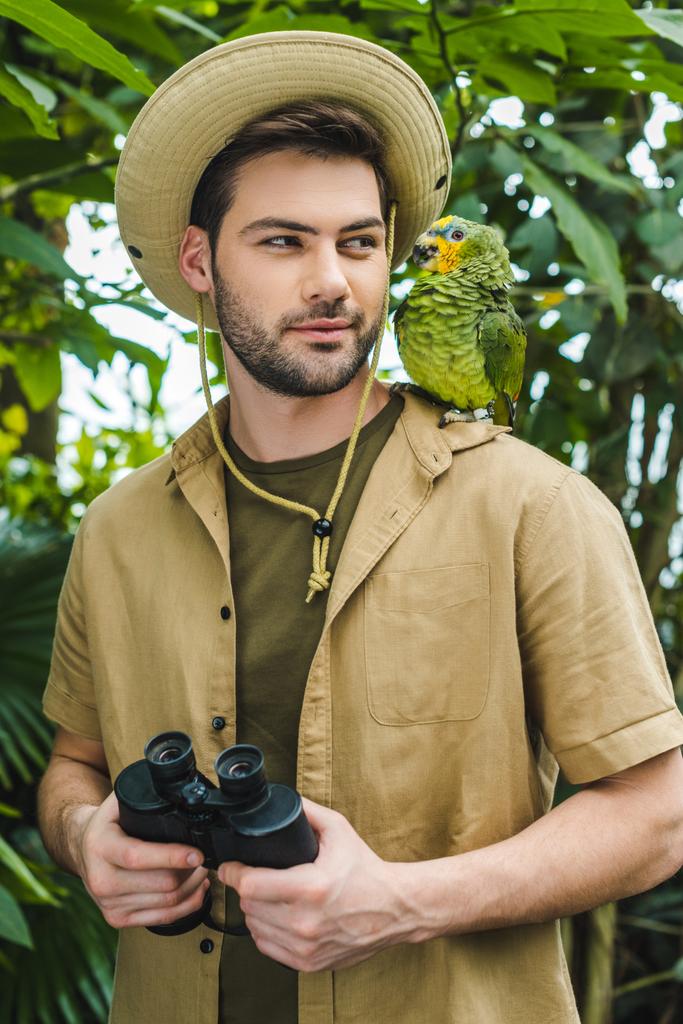przystojny, młody człowiek w kolorze safari ogląda papuga na ramieniu w dżungli - Zdjęcie, obraz