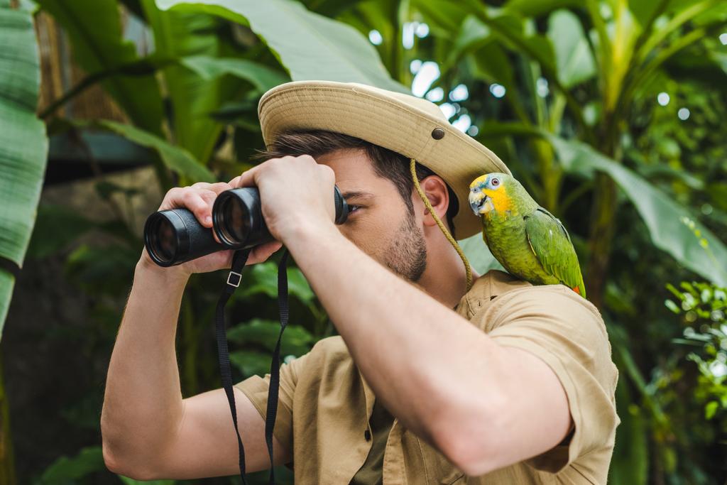 attractive young man with parrot on shoulder looking through binoculars in jungle - Photo, Image