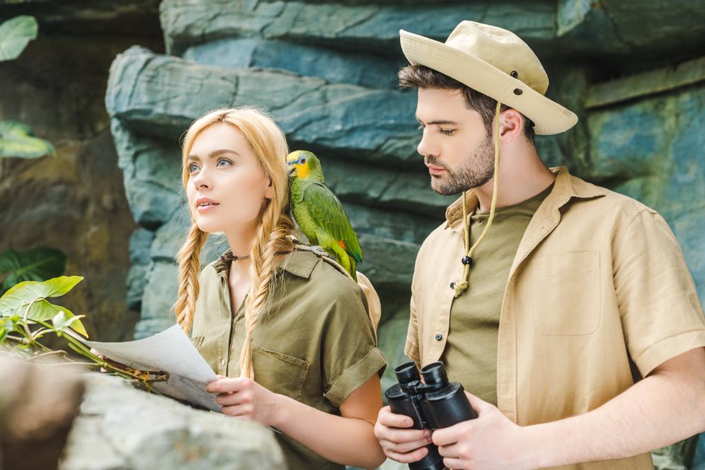 красива молода пара в сафарі костюми з папугою намагається орієнтуватися в джунглях
 - Фото, зображення