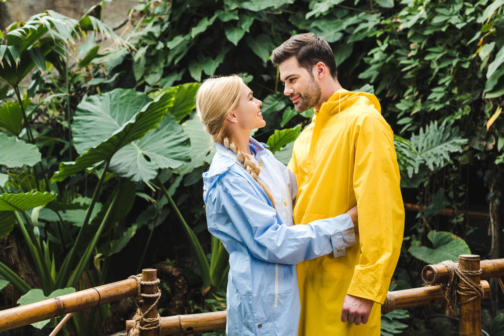 beautiful young couple in raincoats embracing at rainforest - Photo, Image
