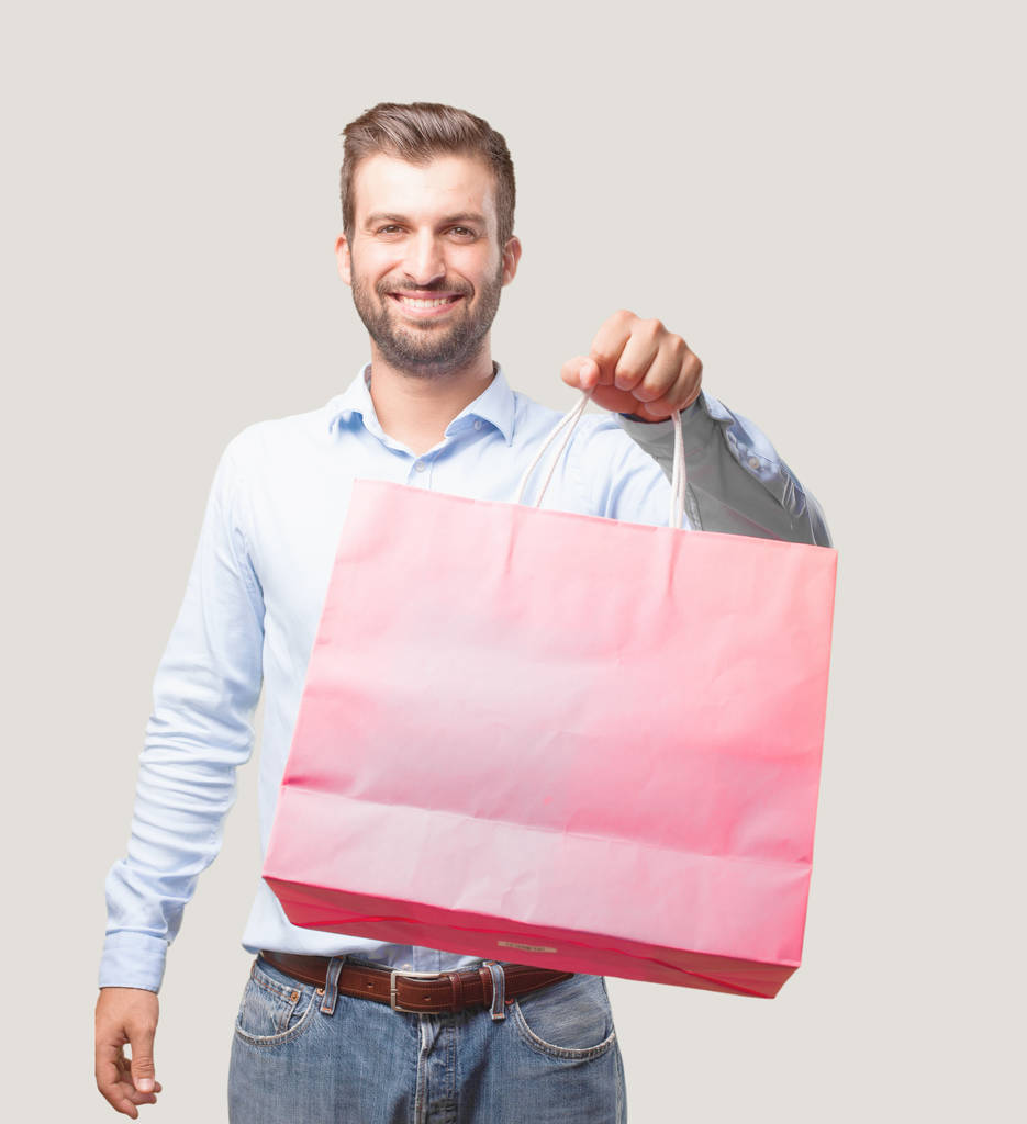 若いハンサムな男、青い t シャツ、ショッピング バッグを押しながら笑みを浮かべてします。幸せなバイヤーのコンセプトです。モノクロの背景に対して隔離される人. - 写真・画像