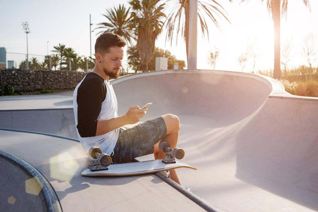 Fiatal ember tart egy mozgatható telefon ülve a skate zóna. Egy srác szörfözhet az interneten a smartphone ülve, a naplemente háttér - Fotó, kép