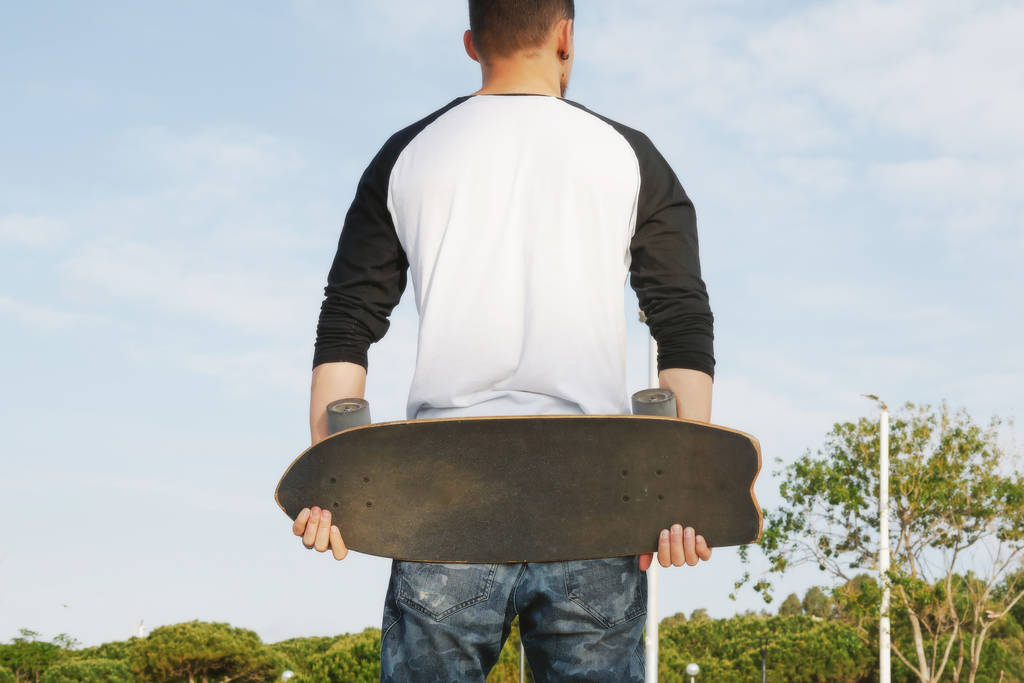 junger Mann in weißem Langarm-T-Shirt steht mit Skateboard im Stadtpark  - Foto, Bild