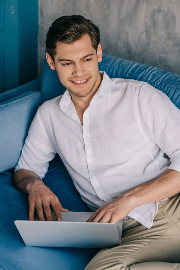 Lächelnder Mann arbeitet auf dem Laptop, während er auf dem Sofa liegt - Foto, Bild