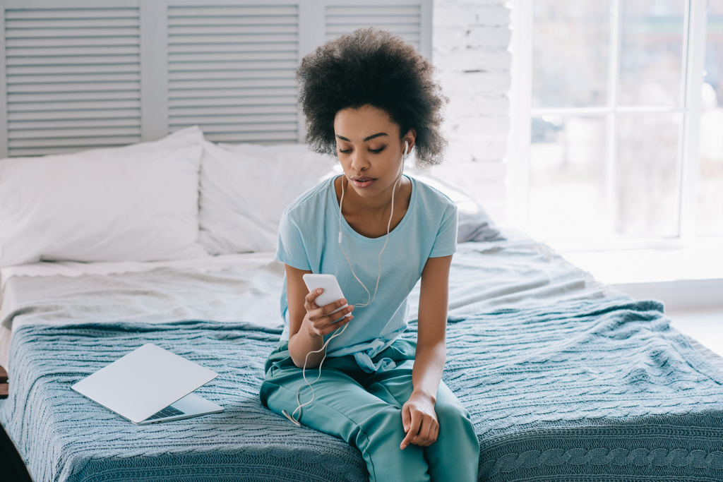 アフリカ系アメリカ人女性のベッドに座って、携帯電話で音楽を聞く - 写真・画像