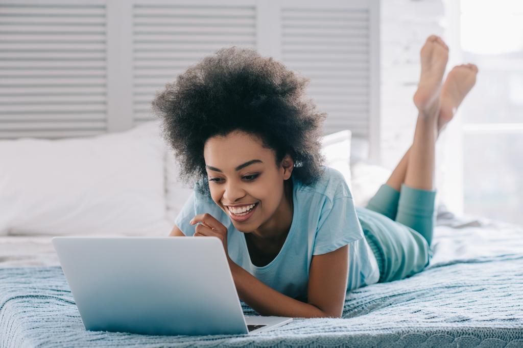 ベッドに横になっていると、ノート パソコンの画面を見て微笑んでいるアフリカ系アメリカ人の女の子 - 写真・画像