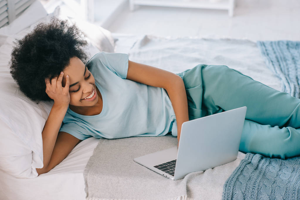 Африканская американка лежит на кровати и смотрит на экран ноутбука
 - Фото, изображение