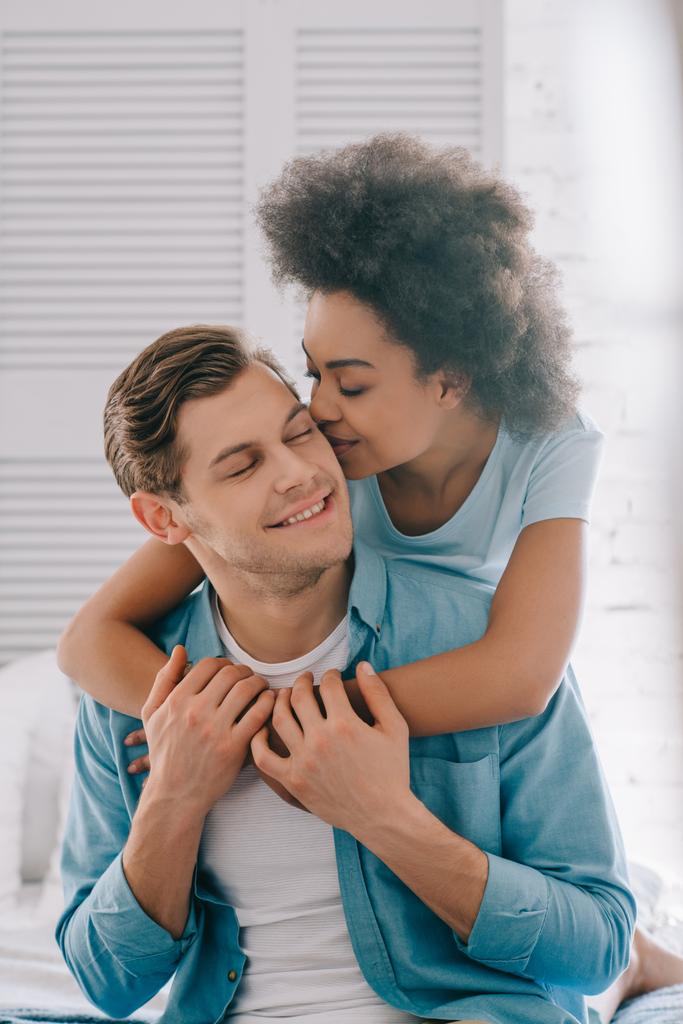 Afrikanische amerikanische Freundin umarmt und küsst Mann, der auf Bett sitzt - Foto, Bild