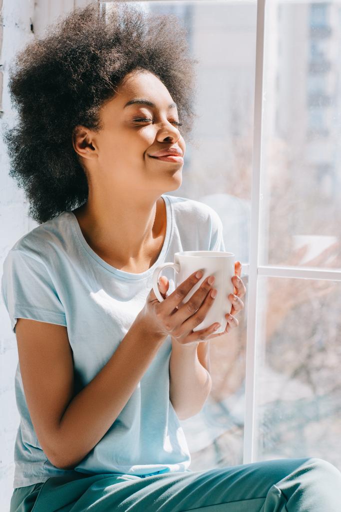 Relajada afroamericana chica sosteniendo taza de café mientras está sentado en el alféizar de la ventana
 - Foto, imagen
