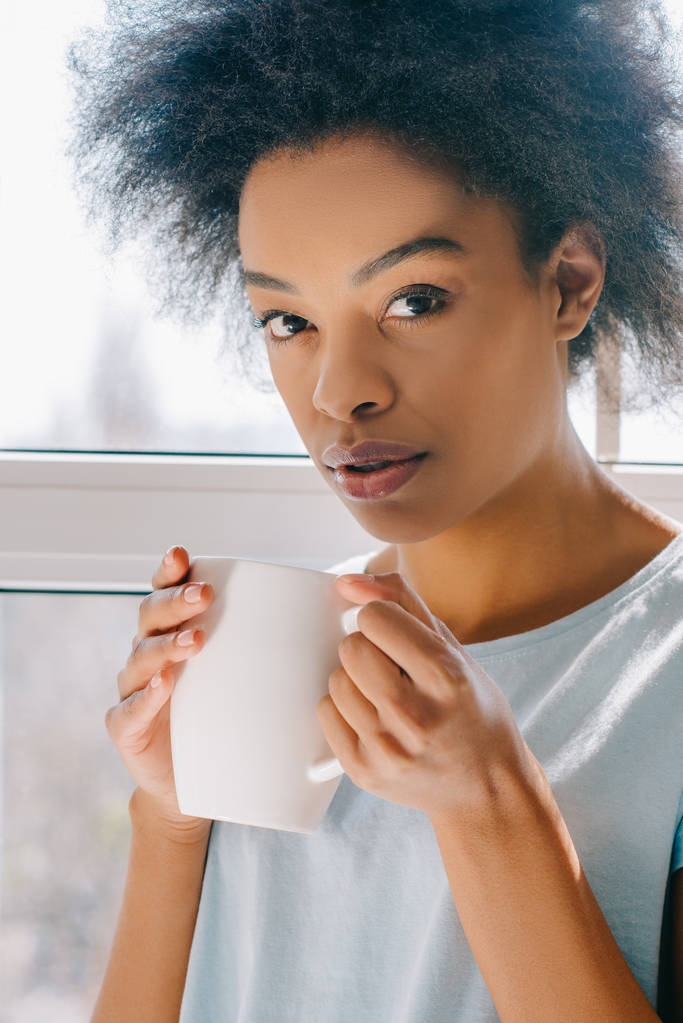 ラウンジでアフリカ系アメリカ人の女の子がカップからコーヒーを飲むを着用します。 - 写真・画像