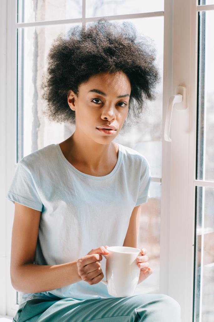 Africká americká žena v obývák oblečení drží šálek kávy zatímco sedí na okenním parapetu - Fotografie, Obrázek