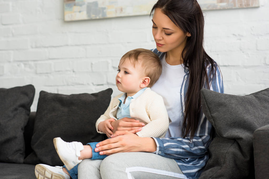 madre con adorable bebé niño en las manos sentado en el sofá en casa
 - Foto, Imagen