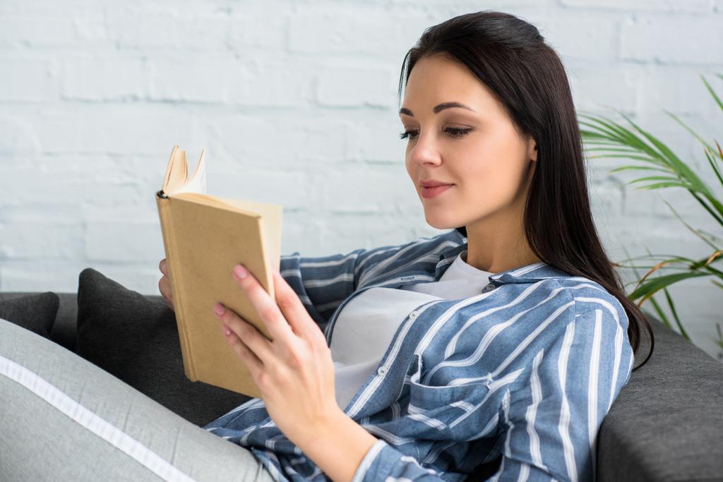 Widok z boku młoda kobieta czytanie książki na kanapie w domu - Zdjęcie, obraz