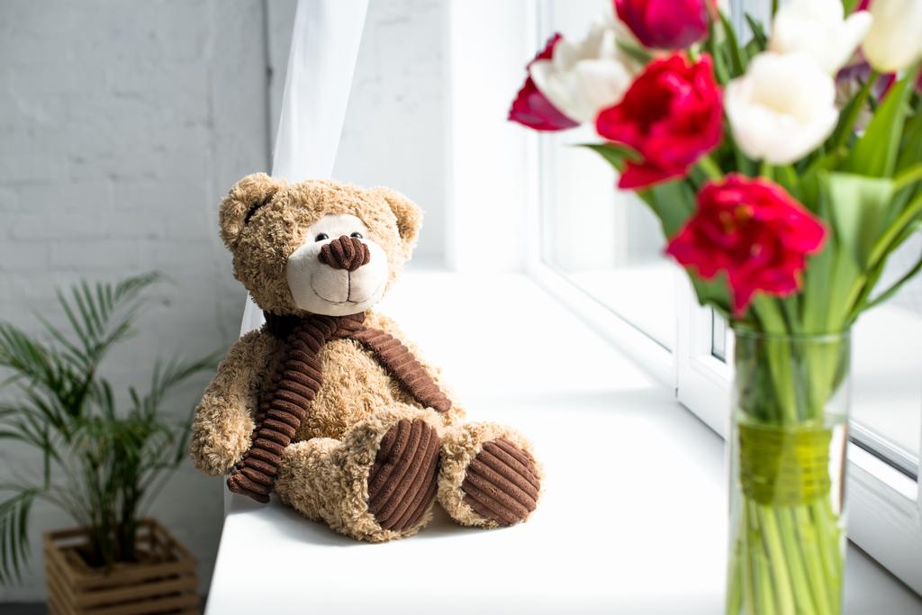 selective focus of teddy bear and bouquet of tulips in vase o window sill at home - Photo, Image