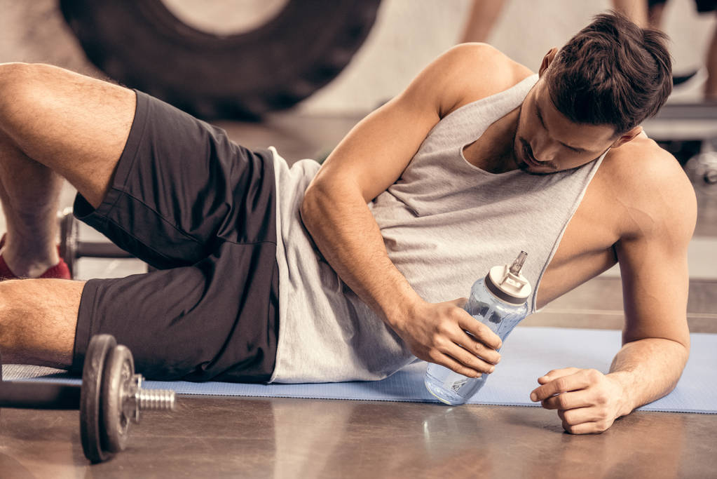 κουρασμένος αθλητή ξαπλωμένος στο πάτωμα σε γυμναστήριο και κρατώντας το μπουκάλι νερό - Φωτογραφία, εικόνα