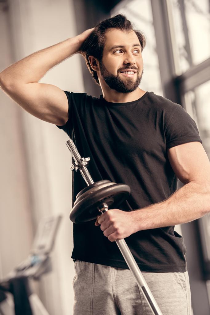 sonriente deportista sosteniendo barra de hierro y tocando el cabello en el gimnasio
 - Foto, Imagen