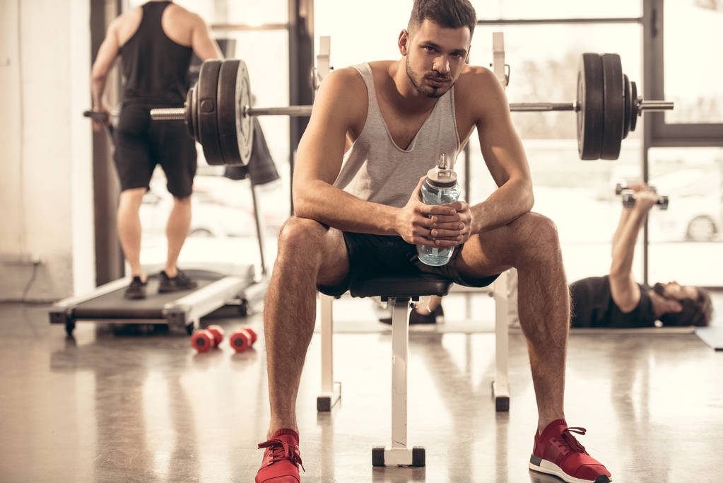 przystojny sportowiec siedzi z butelka wody na wyciskaniu w siłowni - Zdjęcie, obraz