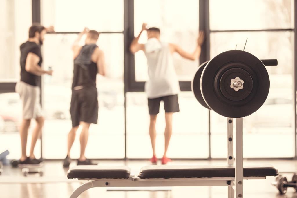 rear view of sportsmen standing near windows in gym with barbell on foreground - Photo, Image