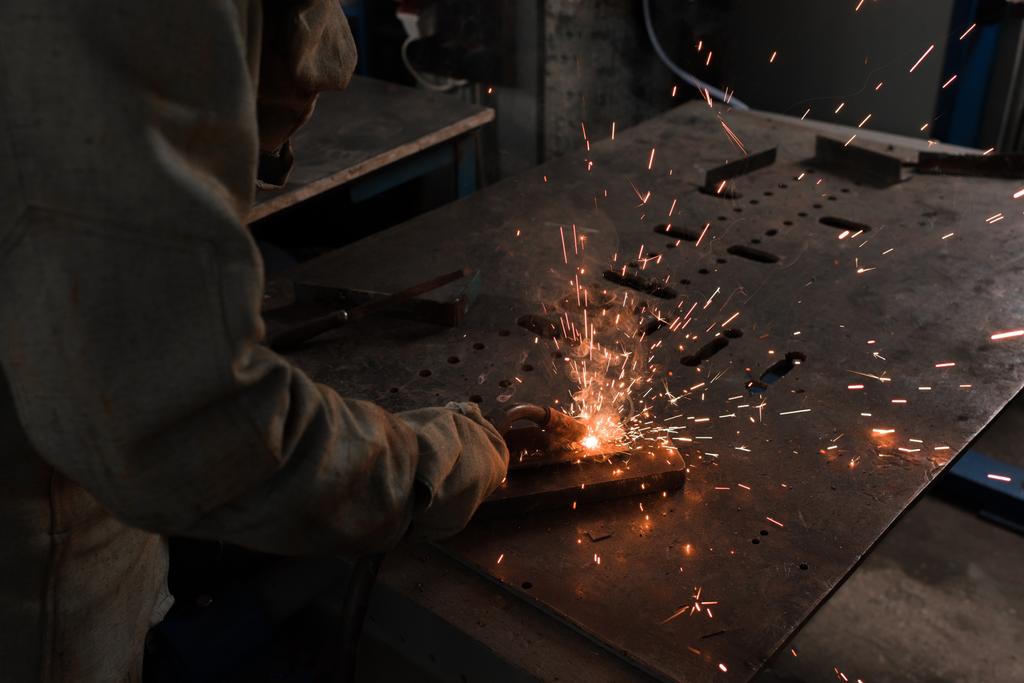 abgeschnittenes Bild eines Arbeiters mit Schutzmaske beim Schweißen von Metall in der Fabrik  - Foto, Bild