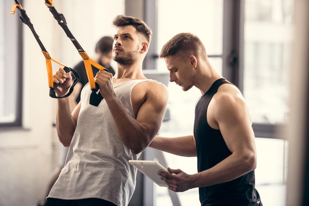 Trainer mit digitalem Tablet und hilft muskulösen Sportlern beim Training mit Fitnessbändern im Fitnessstudio - Foto, Bild