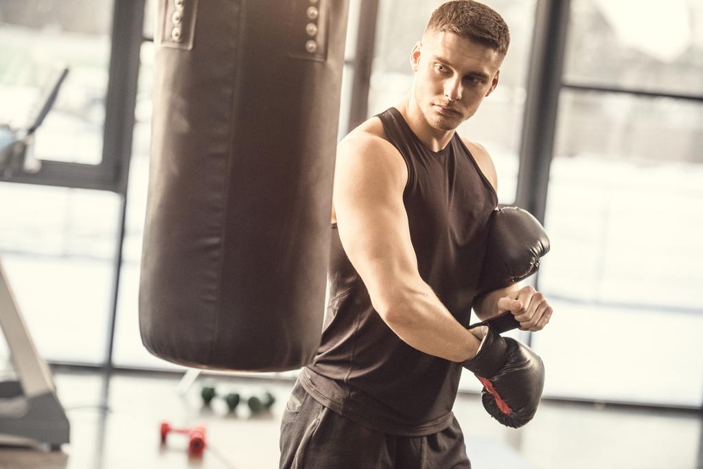 атлетичный молодой человек в спортивной форме в боксёрских перчатках и глядя на боксёрскую грушу в спортзале
 - Фото, изображение