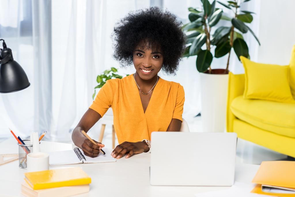 portrait of smiling african american freelancer at workplace with laptop and notebook at home - Photo, Image