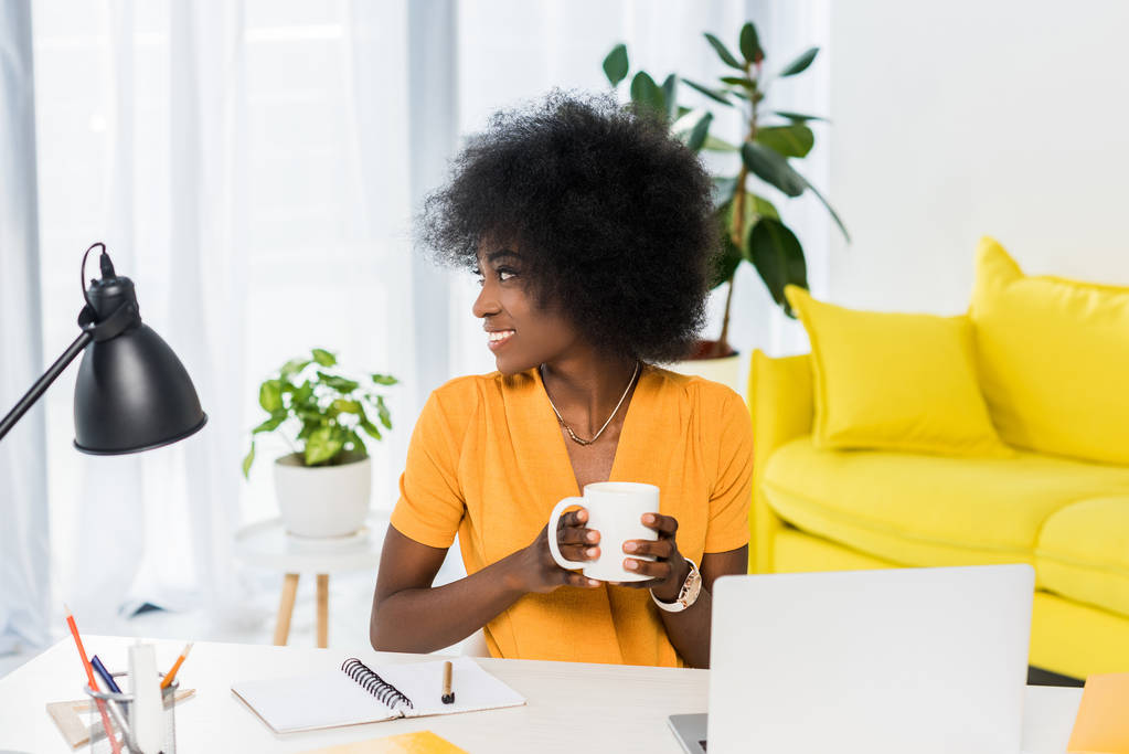 portrait de femme afro-américaine souriante avec une tasse de café dans les mains sur le lieu de travail à la maison
 - Photo, image