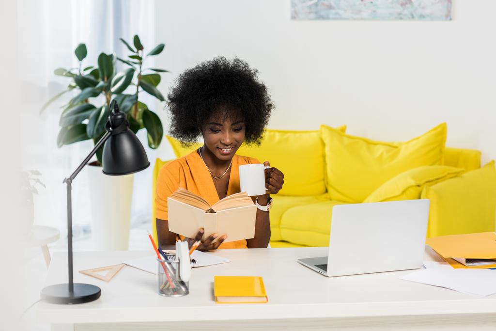 усміхнена афроамериканська жінка з чашкою кави читає книгу на робочому місці з ноутбуком вдома
 - Фото, зображення