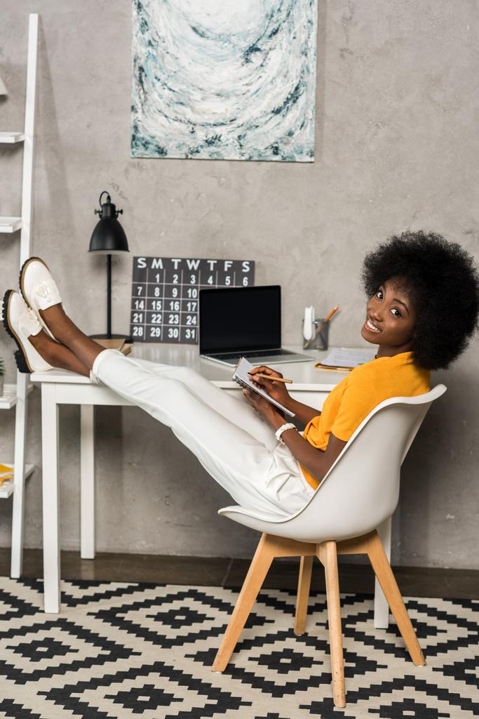 dizüstü bilgisayar vasıl ev ile masada oturma yazıhane defter ile gülümseyen Afrikalı-Amerikalı kadın - Fotoğraf, Görsel