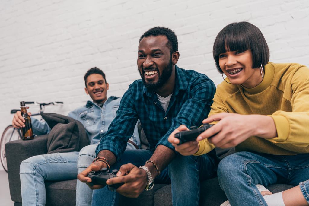 uomo afro-americano sorridente e donna di razza mista con joystick che gioca al videogioco e giovane seduto dietro sul divano con bottiglia di birra
  - Foto, immagini