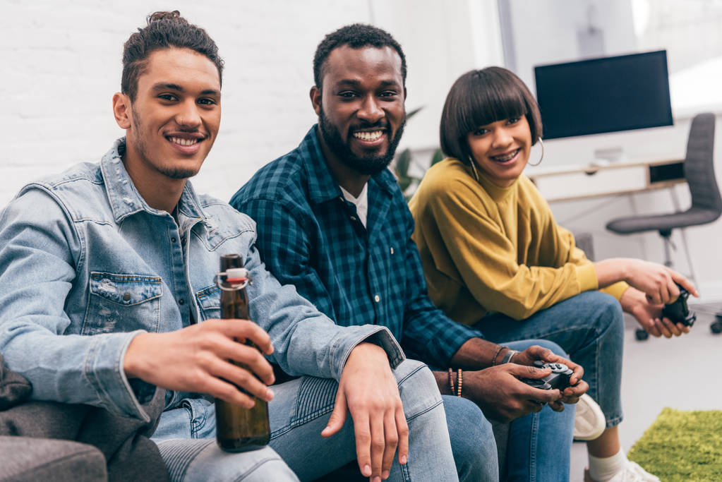 смешанная раса человек с бутылкой пива и улыбающиеся друзья с джойстиками позади
 - Фото, изображение