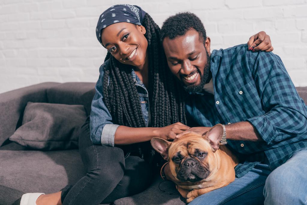 フレンチ ブルドッグとソファの上に座って笑顔の若いアフリカ系アメリカ人のカップル  - 写真・画像