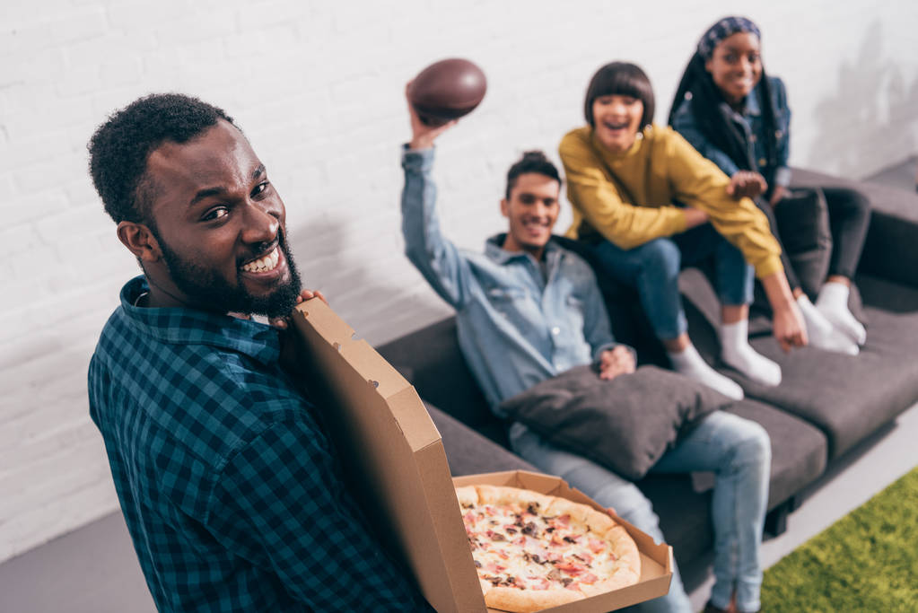 vista ad alto angolo di giovane uomo nero con scatola di pizza e amici sul divano dietro
 - Foto, immagini
