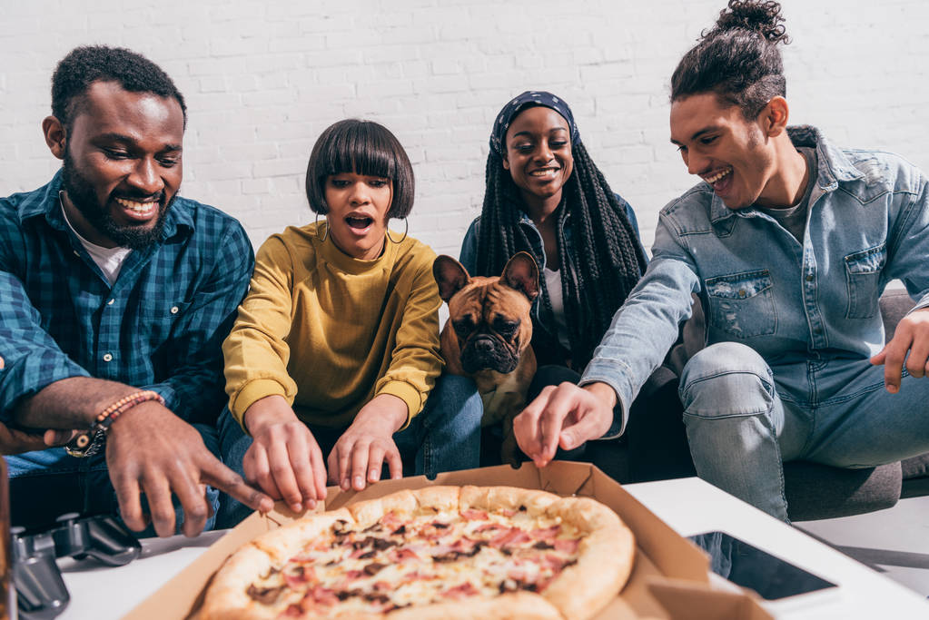 Lächelnde junge multikulturelle Freunde mit französischer Bulldogge beim Pizza essen  - Foto, Bild