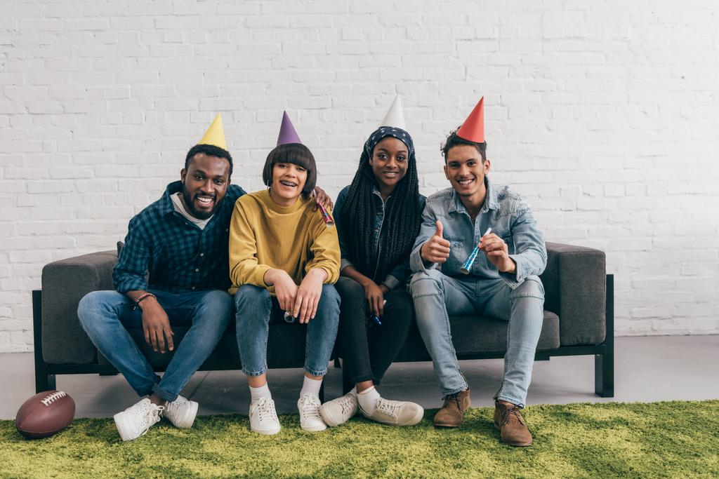 Lächelnde Gruppe junger multiethnischer Freunde in Partyhüten auf der Couch sitzend - Foto, Bild