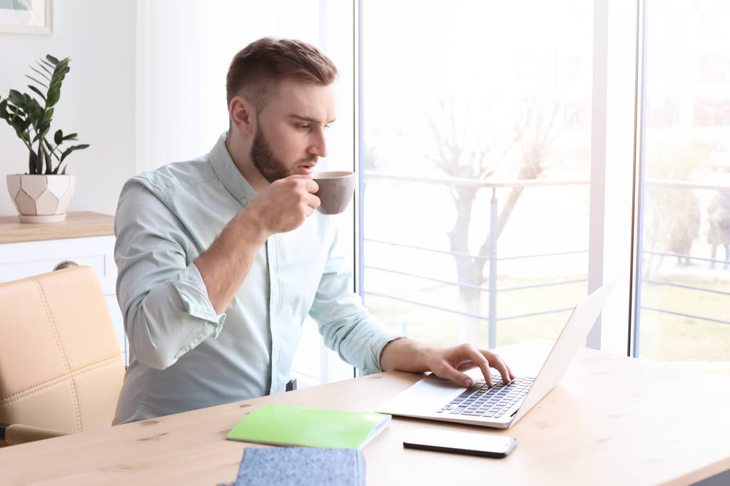 junger Mann trinkt Kaffee, während er mit Laptop am Schreibtisch arbeitet. Heimbüro - Foto, Bild