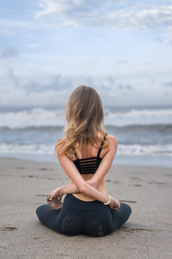rear view of young woman practicing yoga in lotus pose with arms crossed behind back on seashore - Photo, Image