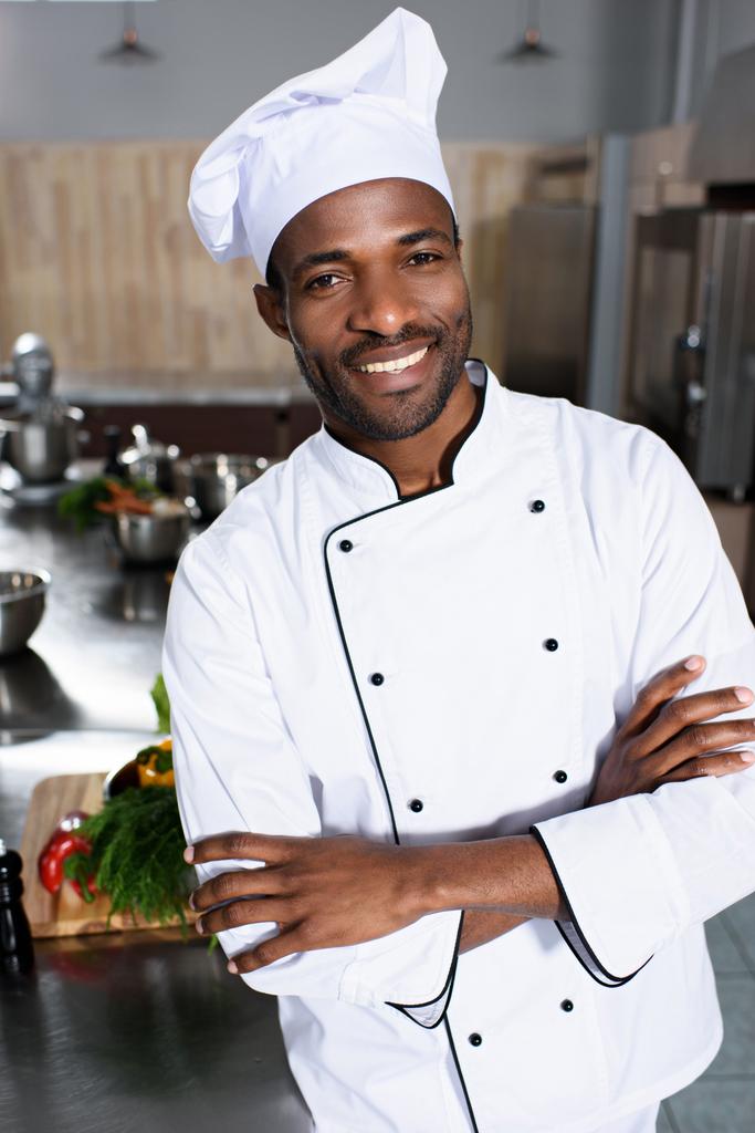 Χαμογελαστά Αφρικανικός Αμερικανός σεφ που στέκεται με τα χέρια διπλωμένα σε σύγχρονη κουζίνα - Φωτογραφία, εικόνα