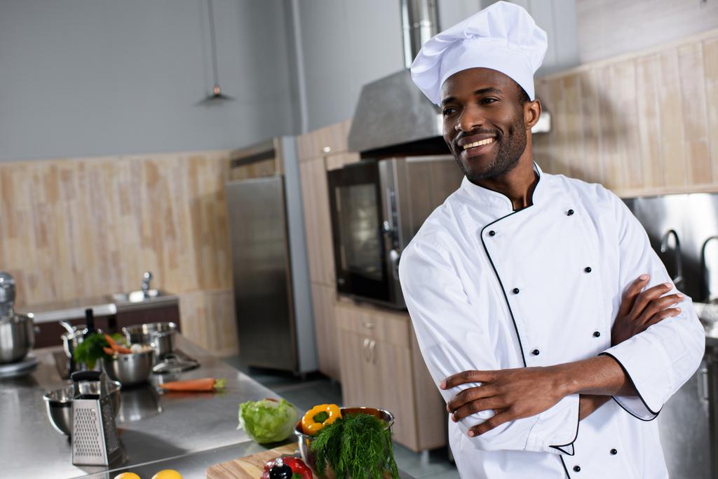Chef afroamericano de pie con los brazos cruzados por la mesa con ingredientes de cocina
 - Foto, Imagen