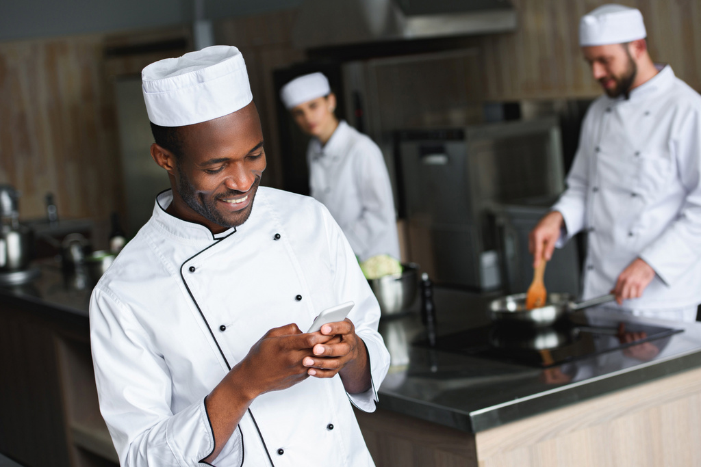 sorrindo chef americano africano usando smartphone na cozinha do restaurante
 - Foto, Imagem