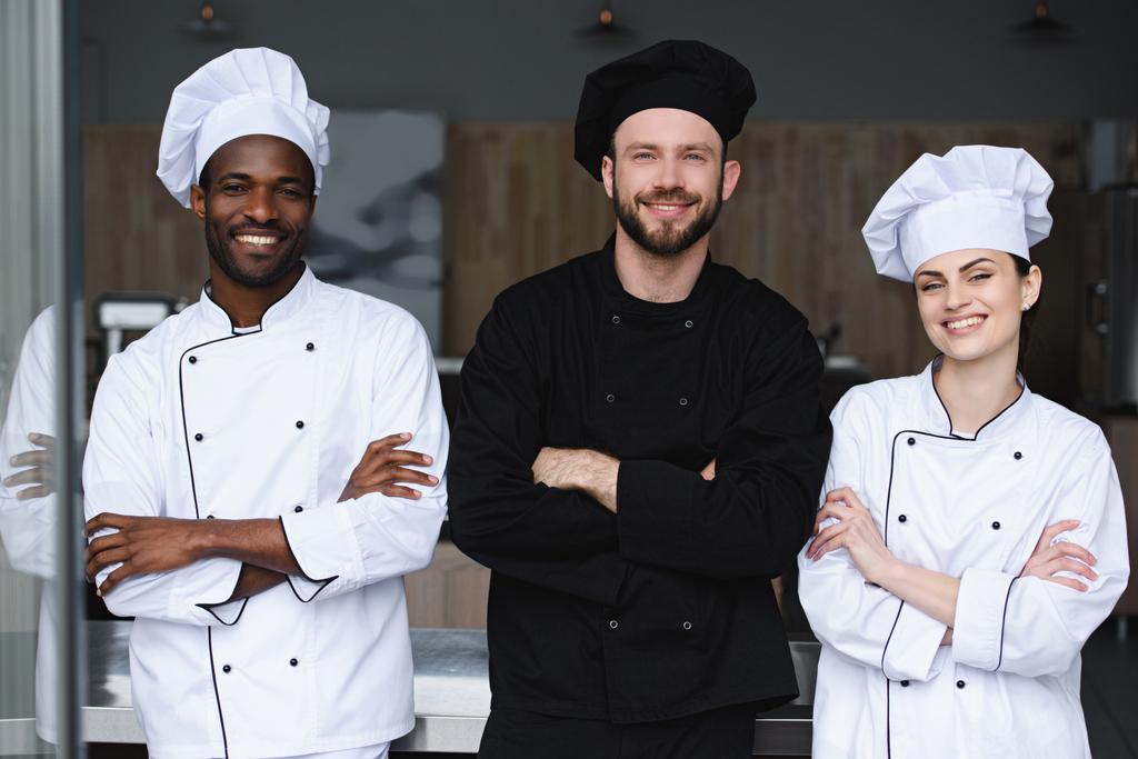 улыбающиеся мультикультурные повара, стоящие со скрещенными руками и смотрящие в камеру на кухне ресторана
 - Фото, изображение