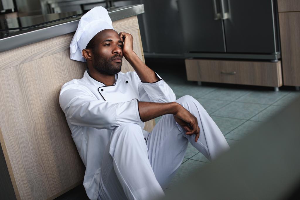 κουρασμένος Αφρικανικός Αμερικανός σεφ κάθεται στο πάτωμα στην κουζίνα εστιατορίου και αναζητούν μακριά - Φωτογραφία, εικόνα