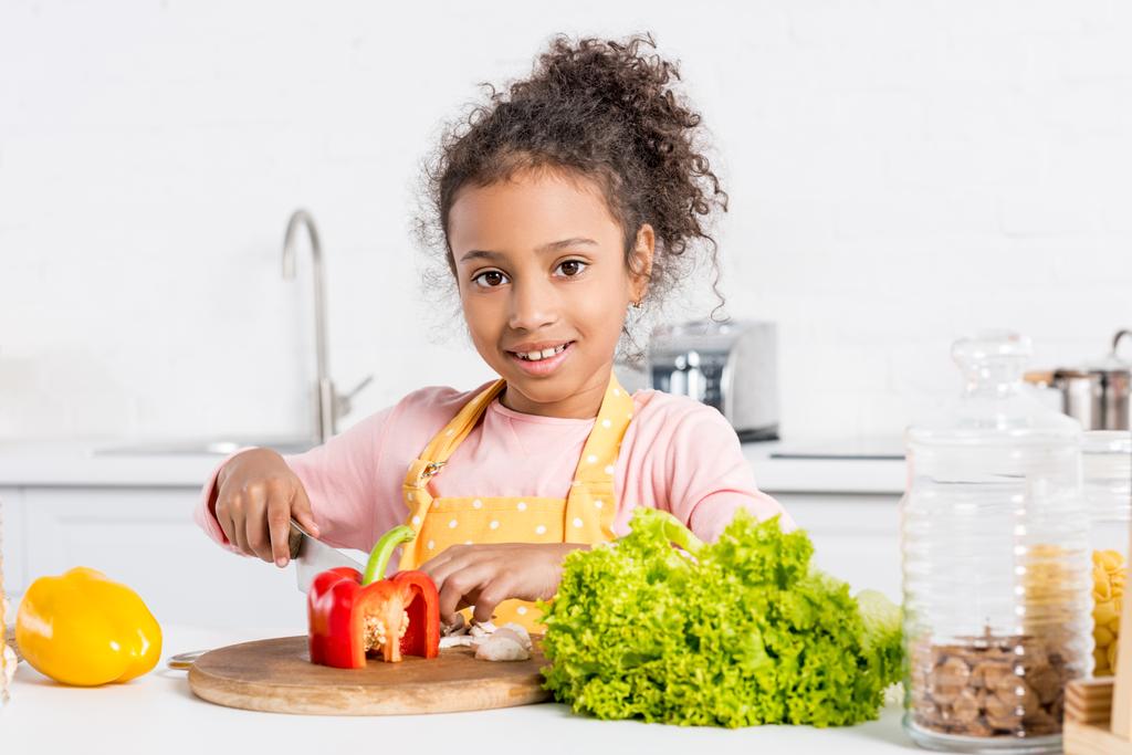 чарівна афроамериканська дитина в фартусі, що ріже болгарський перець і салат на дерев'яній дошці на кухні
 - Фото, зображення