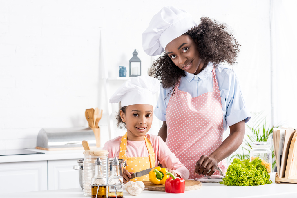 happy african american mother and daughter in chef hats cutting bell pepper on kitchen - Photo, Image